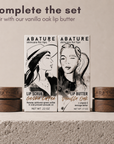 Two ABATURE skincare products displayed on a textured surface: a Lip Scrub Gesha Coffee and a Lip Butter Vanilla Oak, with two matching wooden jars on each side. Text at the top reads 'complete the set. Pair with our vanilla oak lip butter'.
