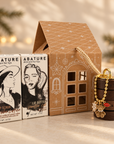 A gift set from ABATURE featuring two lip care products and a decorative house-shaped box. The set includes a Geisha-flavored lip scrub in a white box with black illustration, a Vanilla Lip Butter in a brown box with black illustration, a brown house-shaped container with a gold rope handle, and a small gold charm with a flower and lips detail.