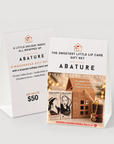 Two white A-frame display cards for Abature Gingerbread Gift Set on a light gray background. The front card shows the product branding, flavor description, and a small holiday-themed illustration with lip balm and a gingerbread houses.