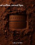 A close-up of a dark brown lip balm product embedded in or pressed into a bed of coffee powder, with the text 'real coffee. unreal lips.