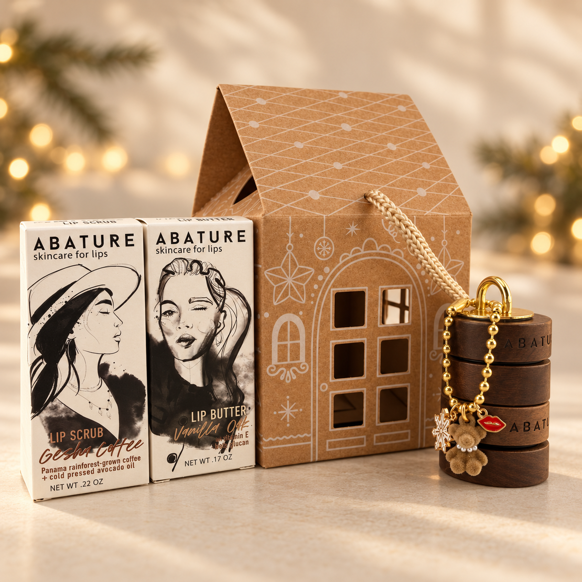 A gift set from ABATURE featuring two lip care products and a decorative house-shaped box. The set includes a Geisha-flavored lip scrub in a white box with black illustration, a Vanilla Lip Butter in a brown box with black illustration, a brown house-shaped container with a gold rope handle, and a small gold charm with a flower and lips detail.