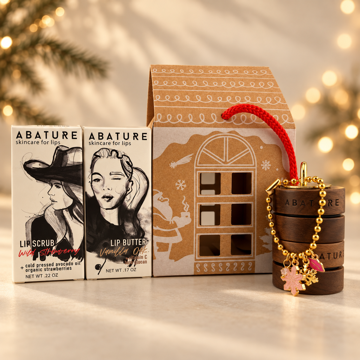 Abature skincare gift set featuring a lip scrub and lip butter box set next to a house-shaped gift box and a wooden stackable jar with a gold beaded bracelet charm, styled with warm holiday lights in the background.