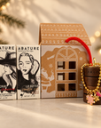 Abature skincare gift set featuring a lip scrub and lip butter box set next to a house-shaped gift box and a wooden stackable jar with a gold beaded bracelet charm, styled with warm holiday lights in the background.