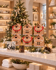 A festive holiday display in a spa-like interior with a decorated Christmas tree, six gingerbread house boxes with red handles, candles, pinecones, and warm shelving in the background. With ABATURE lip balm in the foreground.